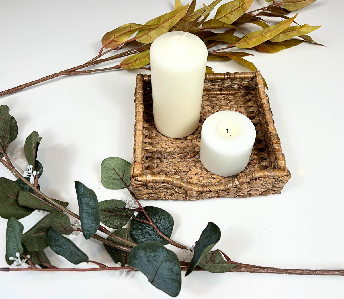 A handwoven beige water hyacinth napkin holder containing two white candles and placed on a white surface with artificial greenery in the background.