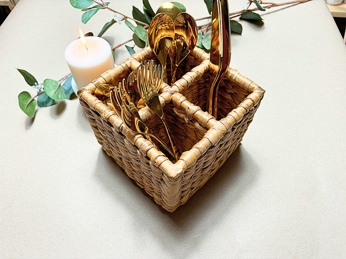 A handwoven natural water hyacinth organizer filled with various kitchen utensils, placed on a table with a candle and decorative leaves in the background.
