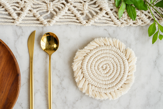 Round hand woven macrame coaster on a marble surface with gold spoon, wooden plate, and macrame wall hanging. Boho dining elegant decor
