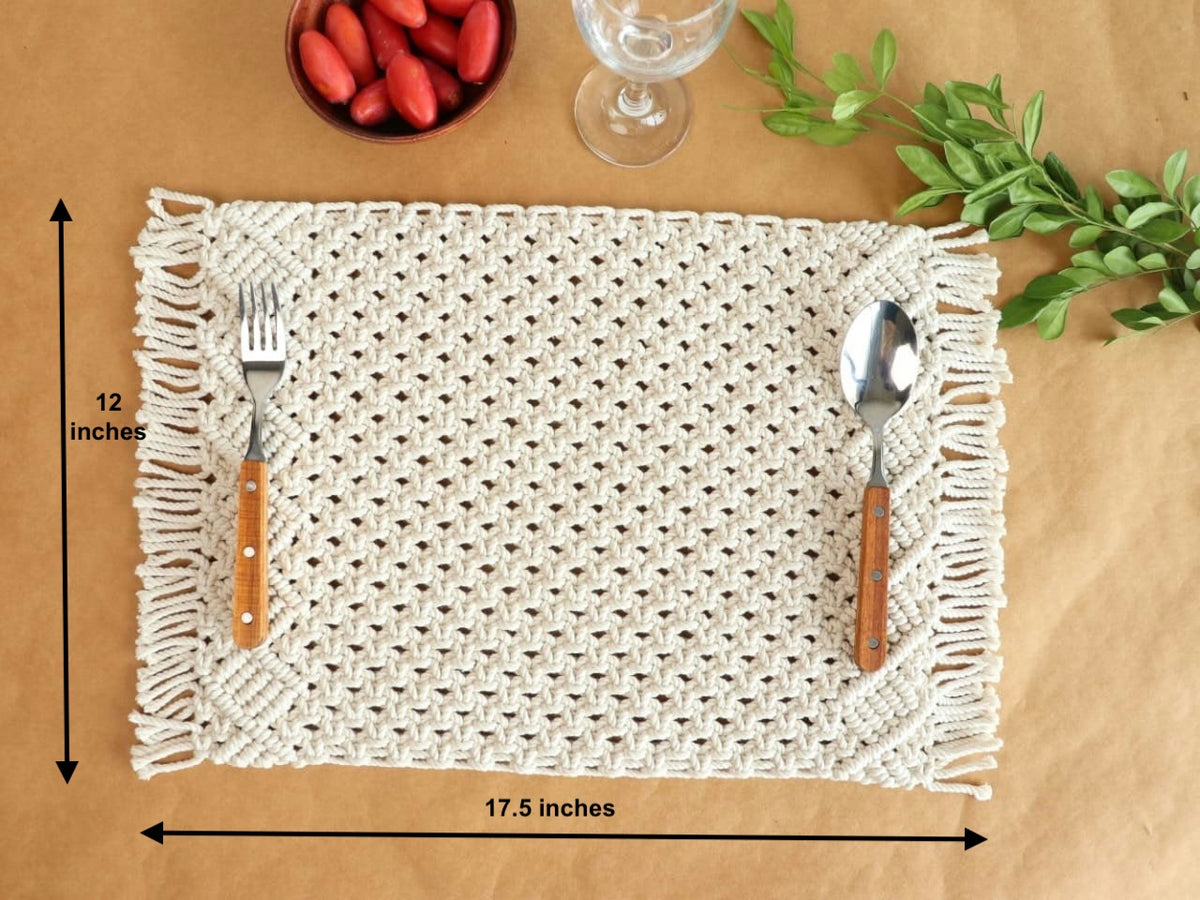 A white boho macramé placemat is displayed on a brown surface with dimensions labeled, alongside a glass, strawberries, and a green plant. The placemat features intricate knots and braided textures.